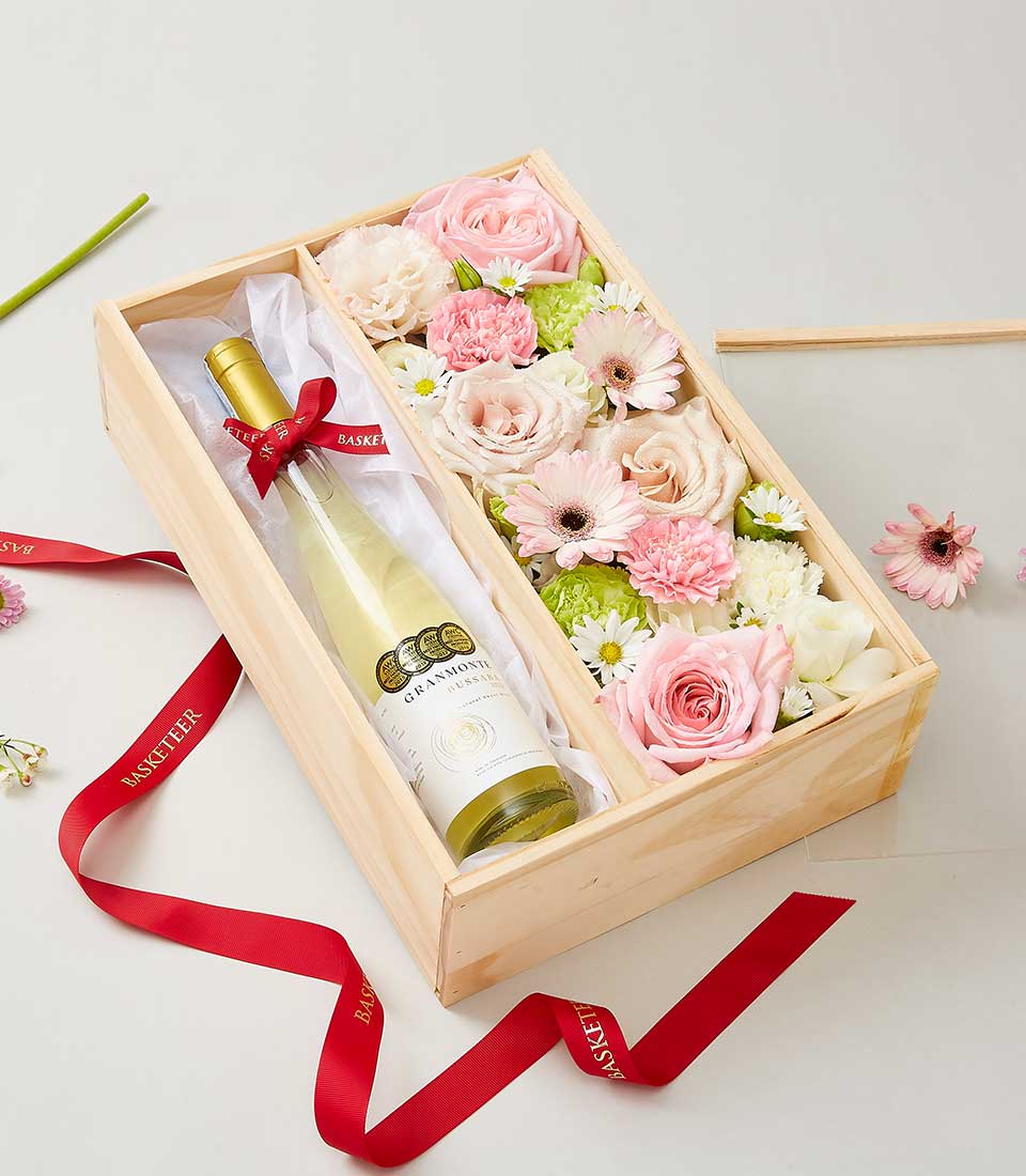 Gift box with a bottle of white wine and a floral arrangement of pink and white flowers in a wooden box.