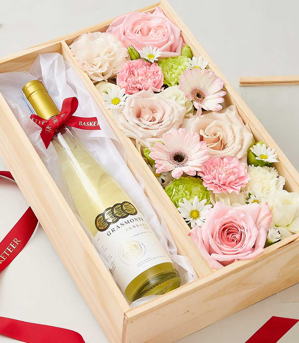 An elegant wooden gift box from Basketeer, showcasing a prestigious bottle of Busaba Sweet Wine, gracefully adorned with a luxurious red ribbon to the left. On the right, an opulent arrangement of pink and white flowers – including exquisite roses, delicate carnations, and charming daisies – meticulously displayed to captivate the senses. Complementing the scene are additional red ribbons that enhance the overall touch of sophistication and charm.