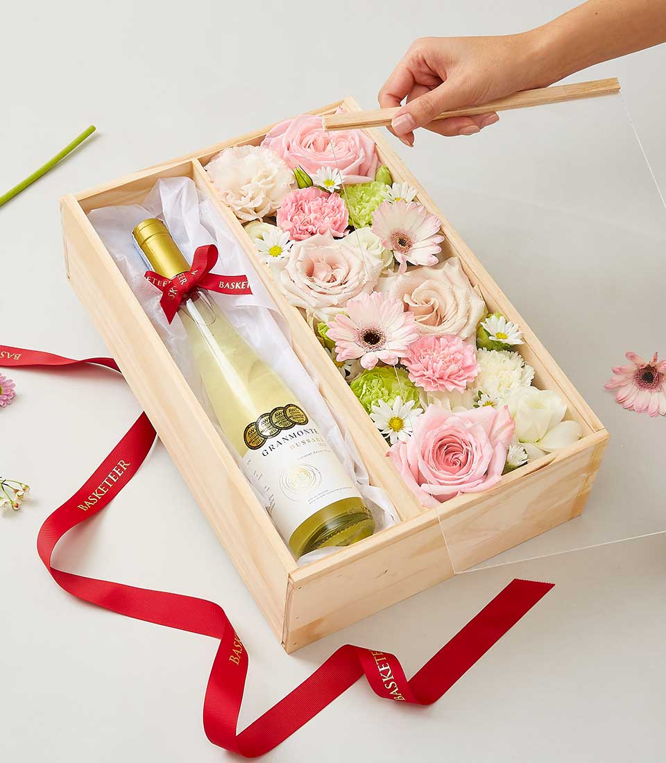 A graceful hand meticulously arranges a delicate pink flower within an elegant wooden gift box, adorned with the prestigious Basketeer logo. To the left, a bottle of exquisite Busaba Sweet Wine nestles perfectly, promising indulgent delight. The box brims with an assortment of luxurious pink and white blooms, including opulent roses and charming carnations, all impeccably arranged to captivate the senses. A sophisticated red ribbon with gold lettering drapes beside the gift box, adding a touch of regal splendor to this magnificent ensemble from Basketeer.