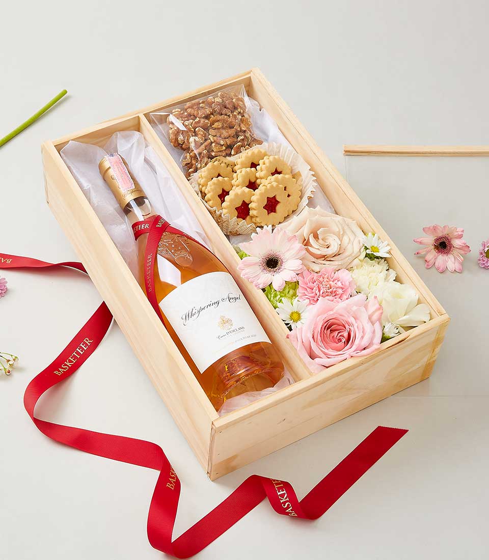 A luxurious wooden gift box from Basketeer elegantly showcases a bottle of exquisite rosé wine, a curated selection of gourmet nuts, and beautifully decorative cookies. Nestled amongst fresh pink roses and daisies, the presentation is further enhanced by red ribbons emblazoned with the Basketeer logo. The backdrop is a serene light neutral canvas sprinkled with delicate flower petals, creating an inviting and sophisticated scene.