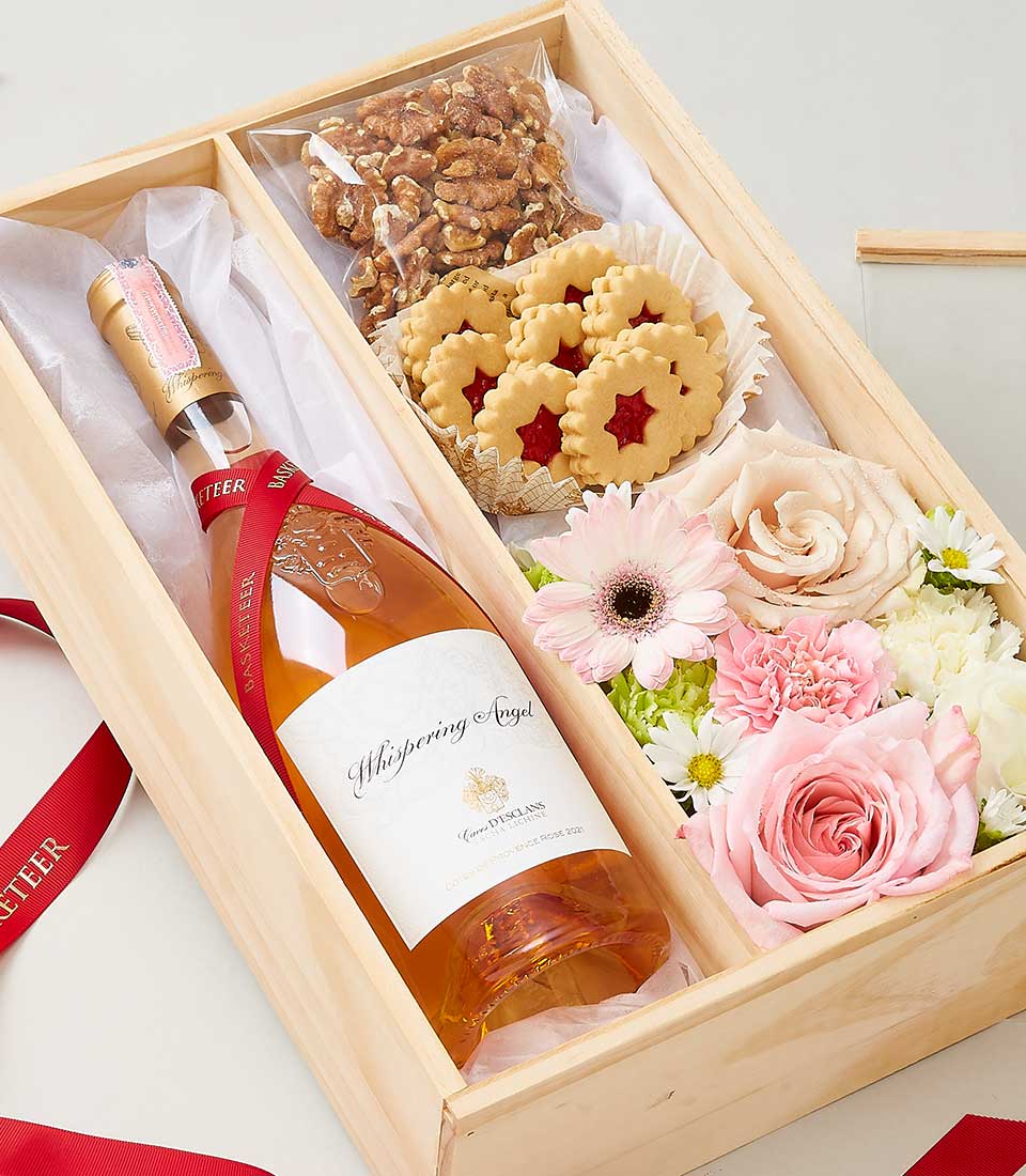A lavish wooden gift box by Basketeer, adorned with an elegant red ribbon bearing the distinguished Basketeer logo, offers a delectable experience. Inside awaits a bottle of exquisite rosé wine, perfectly complemented by a packet of premium shelled walnuts. Star-shaped jam-filled cookies add a touch of sweetness to this refined collection. The arrangement is completed with an opulent selection of pink and white flowers, including delicate roses and vibrant gerbera daisies, all meticulously arranged to create an inviting and luxurious atmosphere.