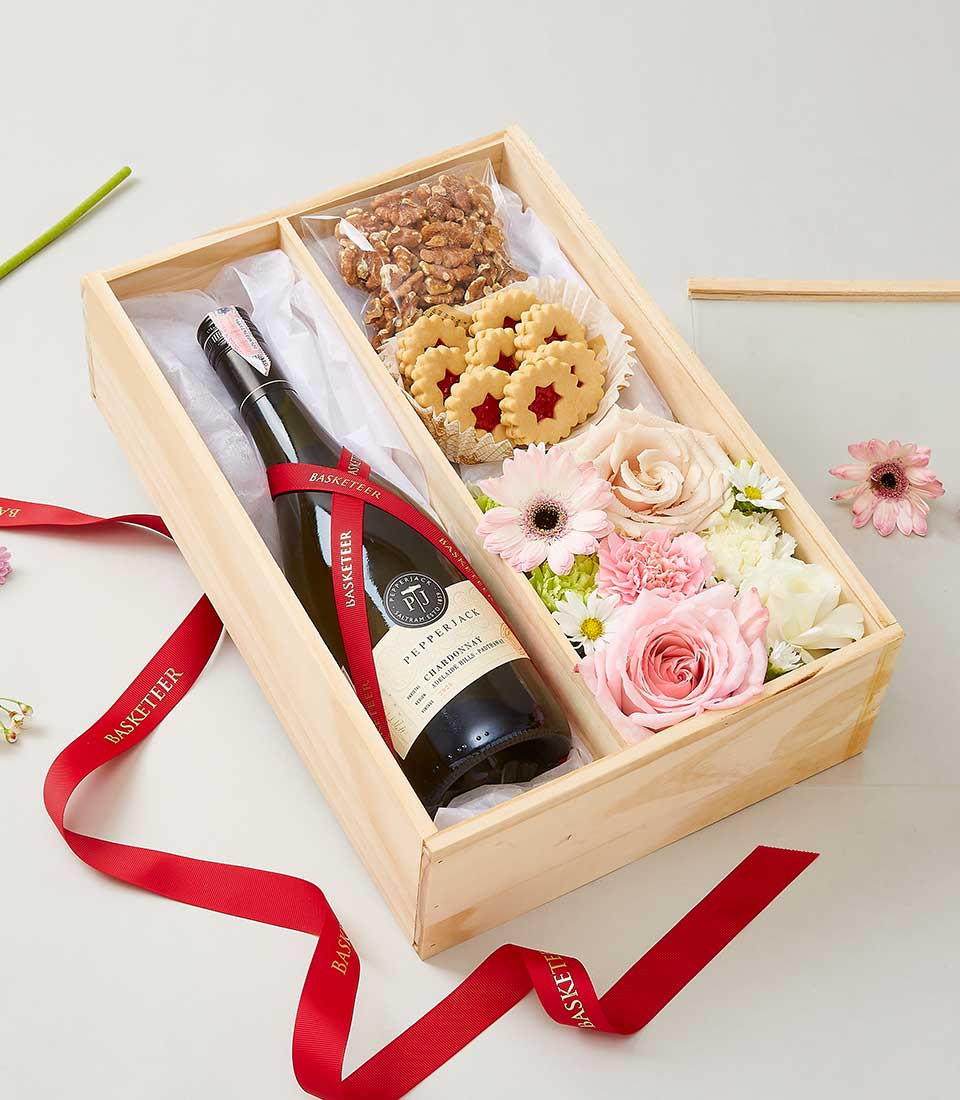 A luxurious wine and cookies gift set from Basketeer features an elegant wooden box containing a premium bottle of red wine, a selection of gourmet assorted nuts, intricately crafted flower-shaped cookies with delectable jam centers, and a vibrant bouquet of fresh flowers including exquisite pink roses and pristine white daisies. Red ribbons elegantly branded with 