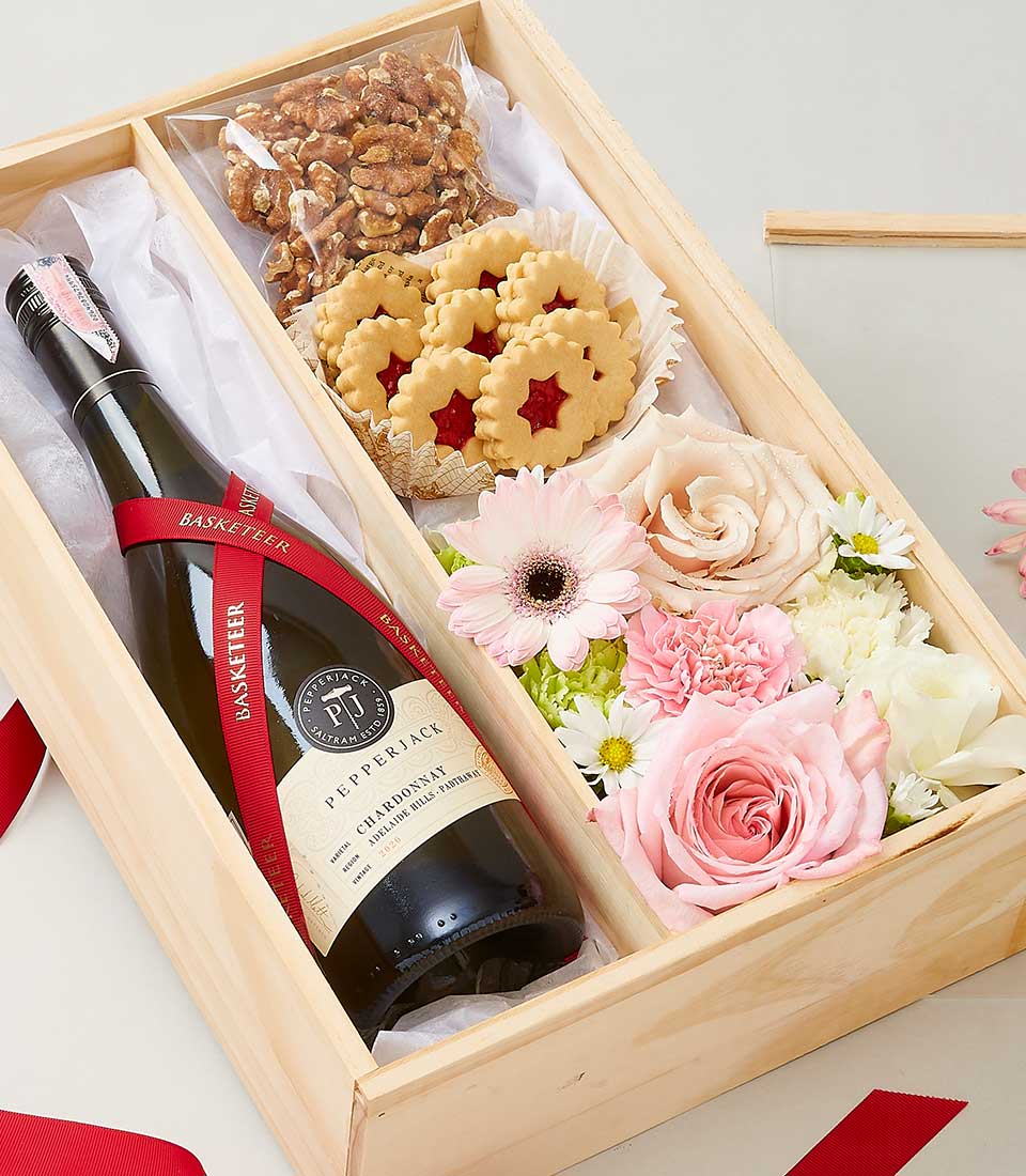 A luxurious and inviting Wine and Cookies Gift Set by Basketeer is gracefully presented in an exquisite wooden box. This elegant collection features a distinguished bottle of Pepperjack wine, complemented by artisanal candied nuts and delectable jam-filled star-shaped cookies. Adding to the allure, a stunning arrangement of pink and white flowers, showcasing radiant roses and cheerful gerberas, enhances the presentation. The box is beautifully adorned with a rich red ribbon, highlighting the exclusivity of this Basketeer creation.