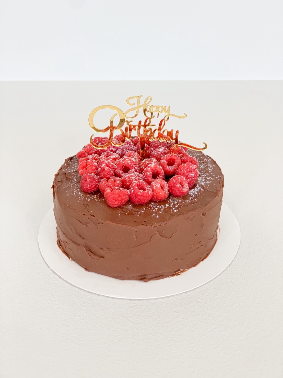 Chocolate birthday cake topped with fresh raspberries and gold happy birthday topper.