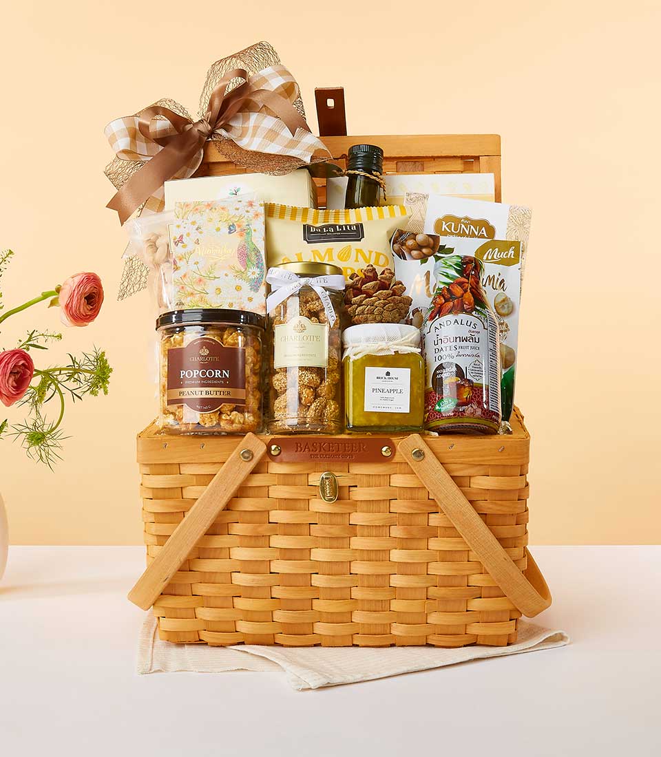 A luxurious picnic basket from Basketeer, artfully arranged with an exquisite selection of gourmet delights: a fine bottle of wine, premium corn kernels, artisanal nuts, delectable snacks, and exquisite jam. The basket is elegantly embellished with a sumptuous ribbon tied in a perfect bow, proudly bearing the Basketeer logo. To the left, a refined vase brimming with fresh flowers complements the scene against a serene peach-toned backdrop. An invitation to indulge in opulence and sophistication.