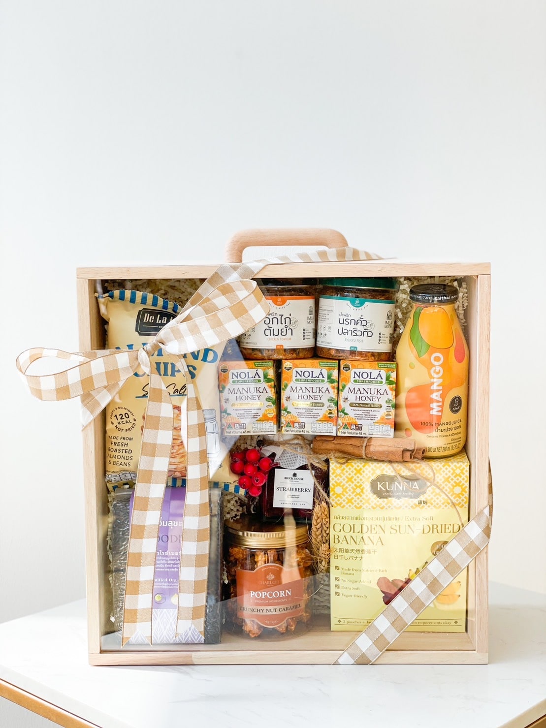 A wooden gift box is filled with assorted snacks and drinks, including jars of spreads, a bottle of mango juice, sun-dried banana slices, a bag of popcorn, and other treats. The box is decorated with a beige plaid ribbon and has a handle on top.