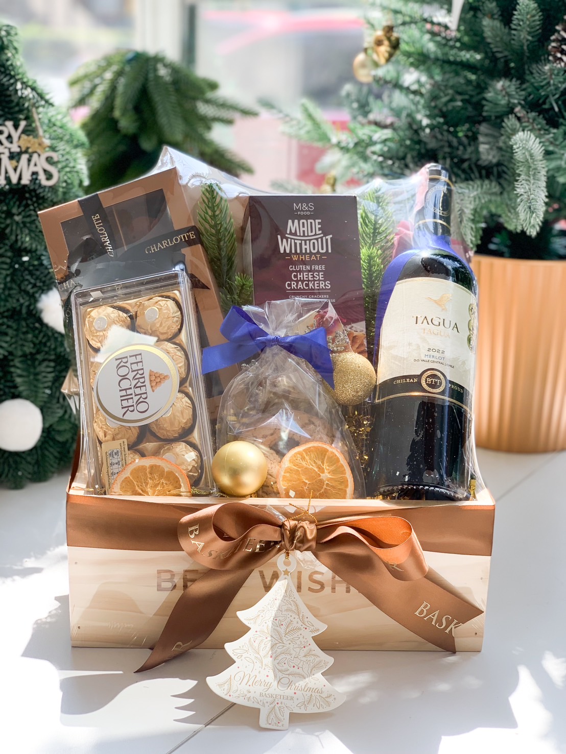 A holiday-themed gift box featuring premium red wine, Ferrero Rocher chocolates, gluten-free crackers, and festive decorations, tied with Basketeer’s signature ribbon.