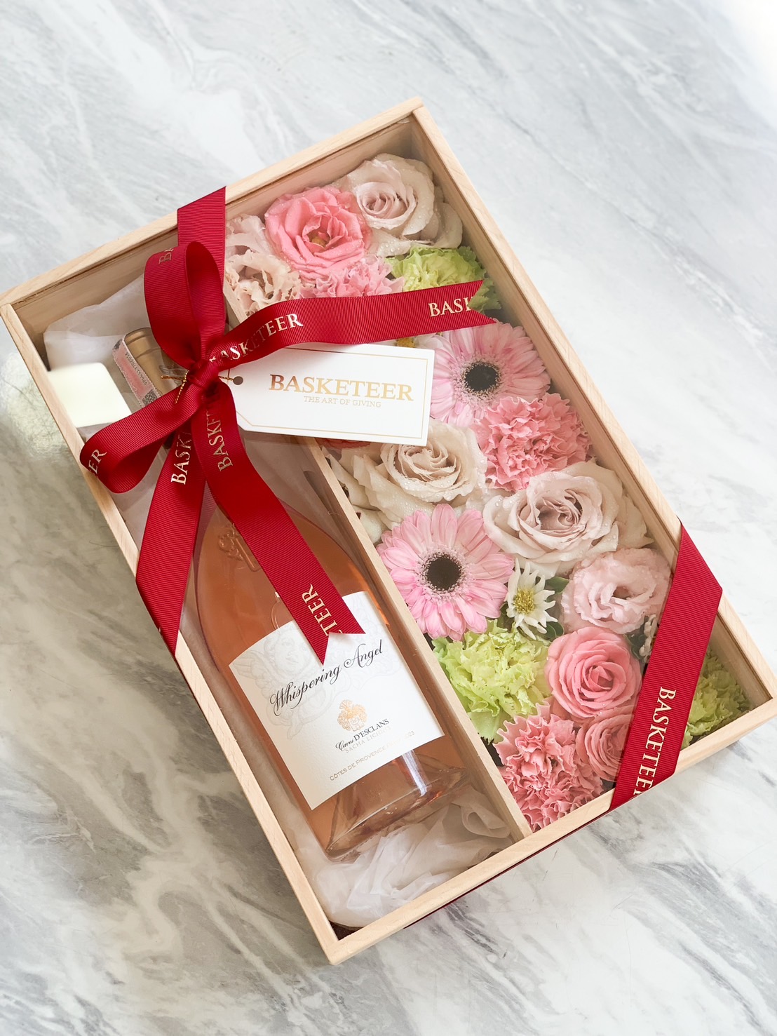 A wooden gift box containing a bottle of Whispering Angel Rosé surrounded by pink and pastel flowers, tied with a red Basketeer ribbon.