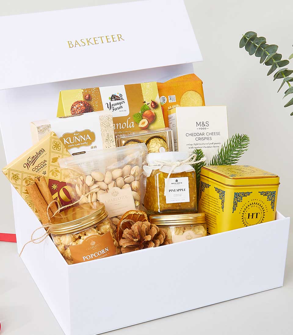 A pristine white gift box adorned with the elegant Basketeer logo reveals an assortment of decadent gourmet delights, including artisanal granola, savory cheddar cheese crispies, premium pistachios, and succulent pineapple in delicate syrup. Luxurious popcorn accompanies a sophisticated tea tin, complemented by gourmet crackers and rich honey. A charming pinecone decoration adds a festive touch, all set against a backdrop of lush greenery, exemplifying opulent indulgence and thoughtful curation by Basketeer.