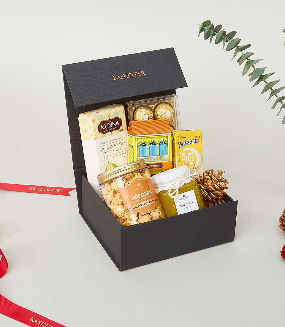 A luxurious Basketeer gift box elegantly adorned with the prestigious Basketeer logo, brimming with gourmet delights including artisanal popcorn, golden honey, buttery biscuits, premium tea blends, and decadent chocolates. The opulent presentation is topped off by a sophisticated red ribbon and a touch of evergreen sprigs for an added festive flair.