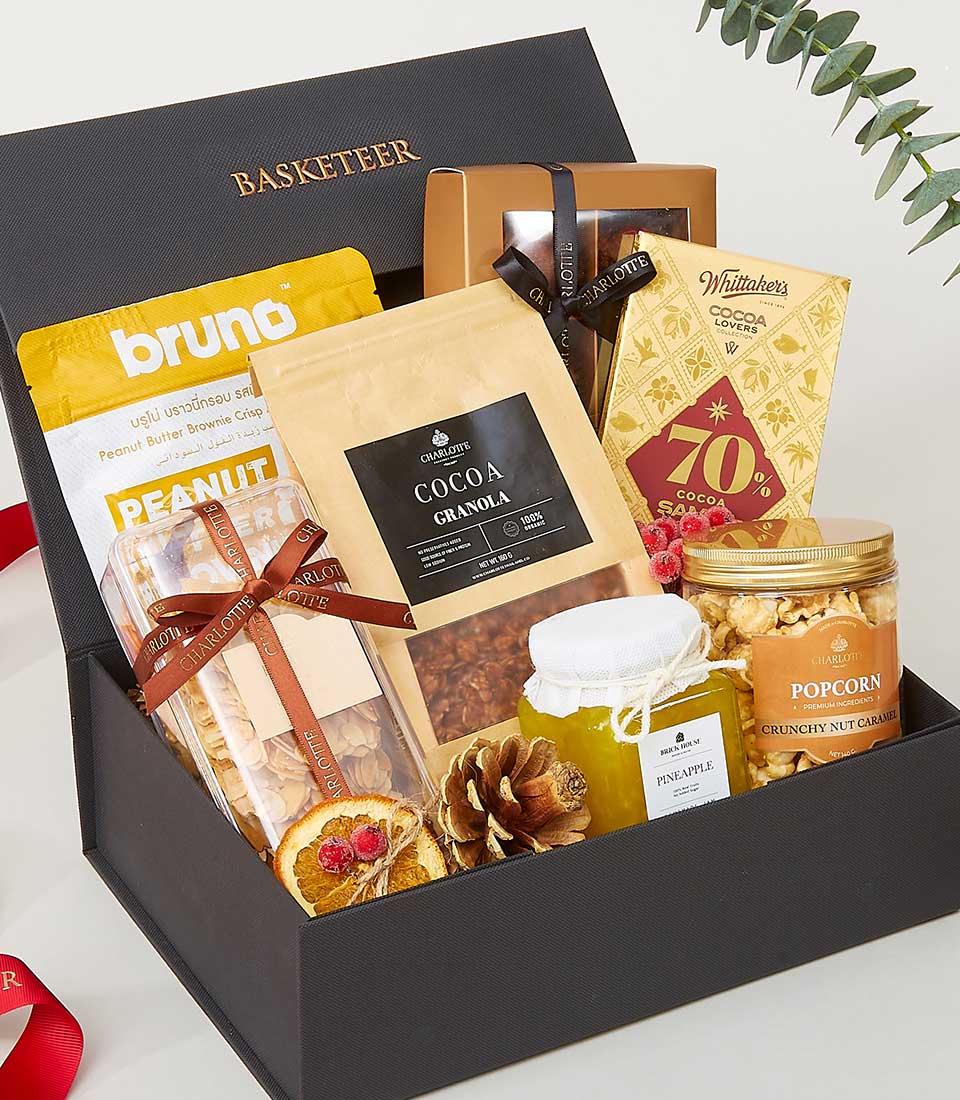 A luxurious black gift box elegantly adorned with the BASKETEER logo showcases an exquisite assortment of gourmet delights. Inside, you'll find rich peanut butter, decadent cocoa granola, artisanal chocolate, premium popcorn, and an indulgent jar of pineapple spread. The opulent presentation is enhanced by elegant ribbons and lush green foliage, complemented by a striking red ribbon that accentuates the sophistication of this lavish gift set.
