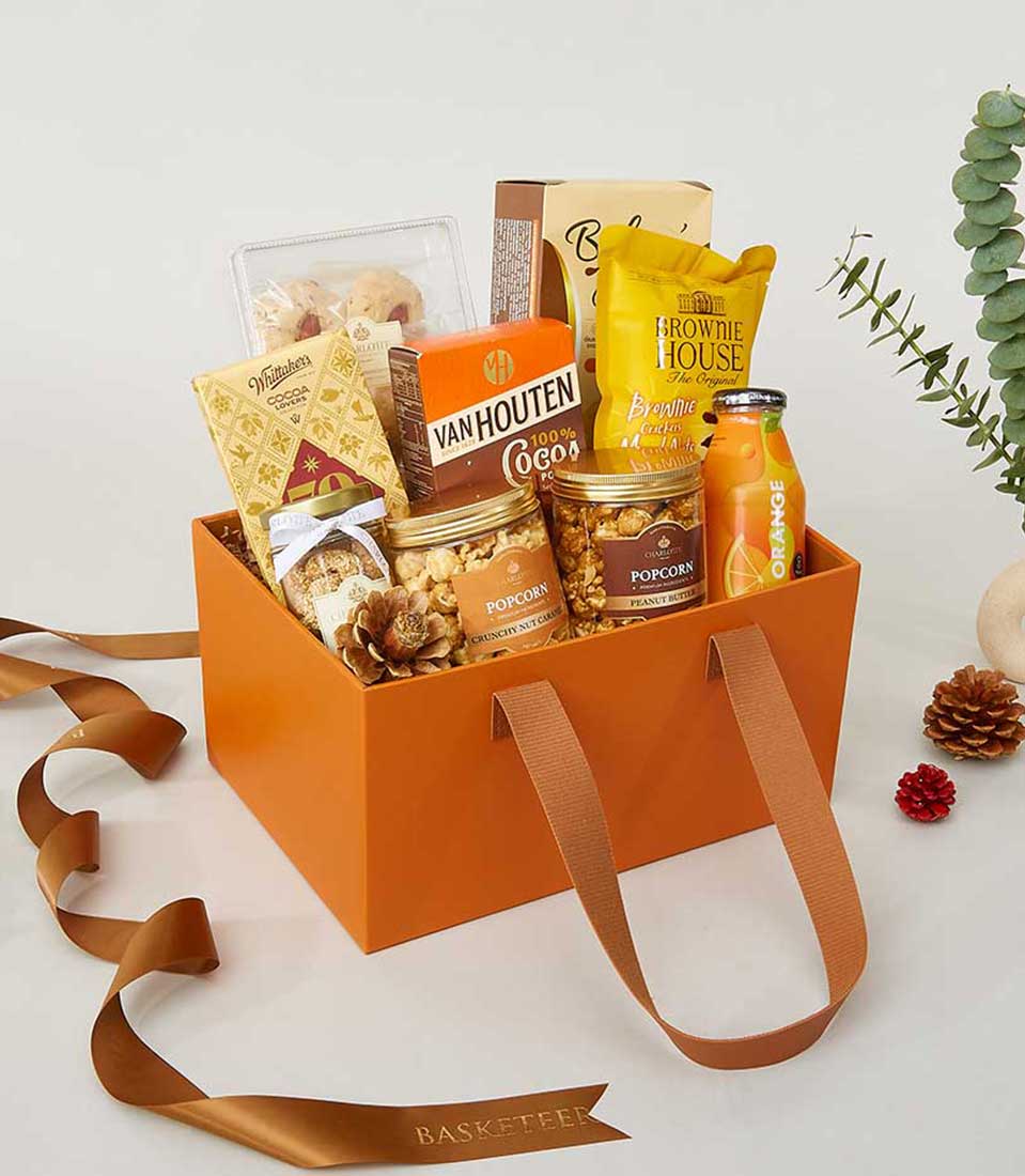 Indulge in the luxurious allure of a Basketeer orange gift basket, elegantly filled with decadent delights. Nestled within this exquisite curation are gourmet popcorn, premium Van Houten cocoa, irresistible brownie crisps, refreshing orange juice, buttery biscuits, rich chocolates, and more culinary treasures. The sophisticated basket features twin handles for easy carrying and is beautifully accented against a pristine white backdrop adorned with festive pinecones and lush green foliage. A touch of elegance is added by the brown ribbon elegantly bearing the distinguished Basketeer logo.