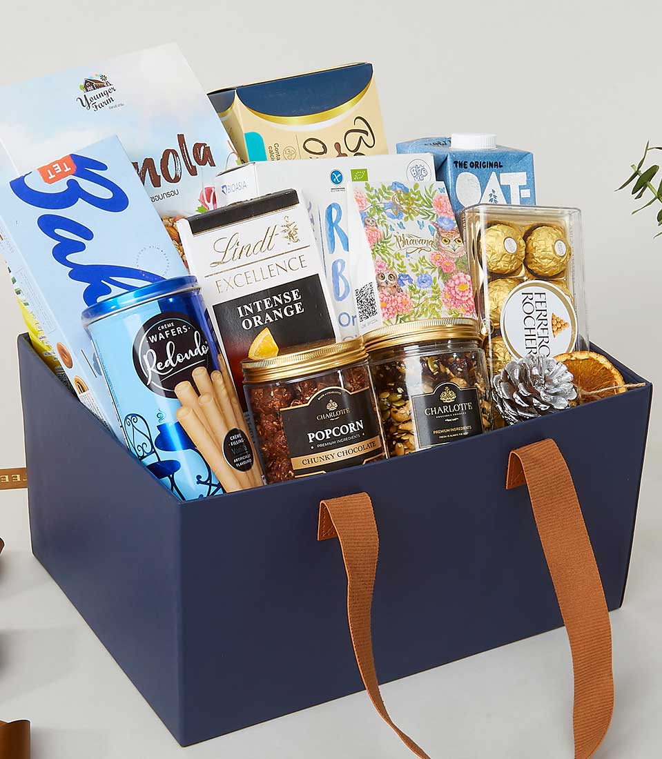 A sumptuous blue gift box adorned with sophisticated brown handles, lavishly filled with an array of gourmet delights. Indulge in the Basketeer experience with artisanal granola, exquisite Lindt Excellence Intense Orange chocolate, handcrafted popcorn, soothing herbal tea blends, savory crackers, decadent Ferrero Rocher chocolates, and a petite jar of golden honey. A charming pine cone adds a touch of festive elegance to this inviting ensemble. The tasteful packaging prominently features the prestigious Basketeer logo on a ribbon and box, ensuring an unparalleled gifting presentation.