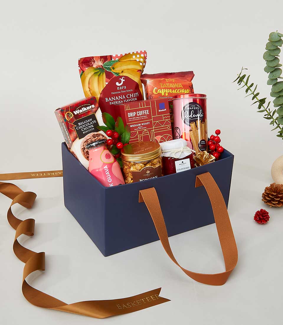A luxurious navy blue gift basket with elegant brown handles, overflowing with an exquisite selection of gourmet snacks and beverages from Basketeer. Delight in the assortment of banana chips, rich cappuccino mix, artisanal drip coffee, decadent cookies, and premium honey and jam. The sophisticated arrangement is set amidst festive decorations, lush greenery, and a tastefully adorned ribbon featuring the distinctive Basketeer logo.