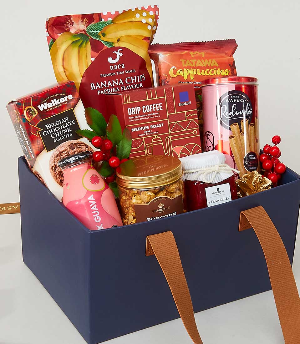 A luxurious navy gift box from Basketeer, brimming with an exquisite assortment of snacks and treats. The presentation includes sophisticated paprika-flavored banana chips, a rich cappuccino mix, tantalizing strawberry jam, decadent chocolate wafer rolls, gourmet Belgian chocolate biscuits, artisanal popcorn, a refined medium roast drip coffee selection, and a refreshing bottle of guava juice. Adorned with festive red berries and lush green leaves to elevate the look even further. A perfect embodiment of elegance and indulgence by Basketeer.
