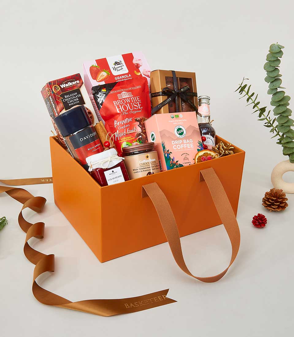 An opulent orange gift box elegantly featuring the distinguished Basketeer logo and ribbon, boasting two graceful handles. This luxurious assortment includes an array of gourmet delights: a fine bottle of drink, artisan drip bag coffee, delectable jam, wholesome granola, premium brownie mix, and other exquisite treats. Enveloping this splendid ensemble are festive adornments like lush green leaves, rustic pine cones, and chic ribbons—creating an enchanting and inviting atmosphere.