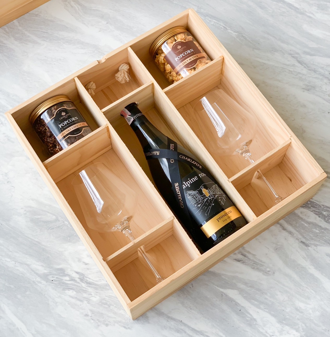 A wooden gift box with compartments containing a bottle of red wine, two wine glasses, and two jars of gourmet popcorn. The items are neatly arranged on a light, marbled surface, making for the perfect Exclusive Date Night Wine Collection.