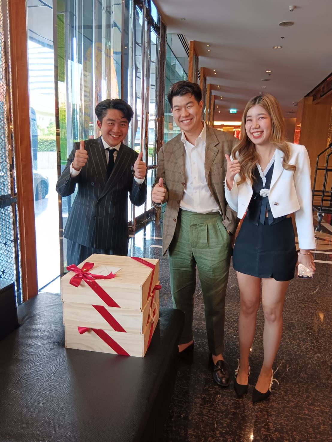 Three people stand indoors next to a table with wooden gift boxes wrapped in red ribbons. The leftmost person is in a striped suit, the middle in a plaid blazer and green pants, and the rightmost in a black skirt and white blazer. They're all smiling and giving thumbs up, celebrating their Exclusive Date Night Wine Collection.