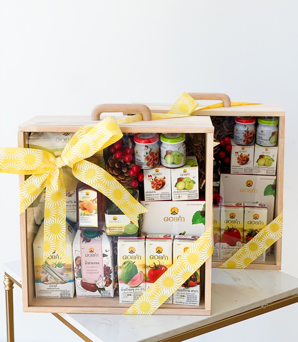 A gift set featuring two wooden boxes filled with various beverage bottles and boxed drinks is adorned with yellow ribbons and decorated with red berries and pinecones, placed on a white and gold table. The wooden boxes have handles for easy carrying.
