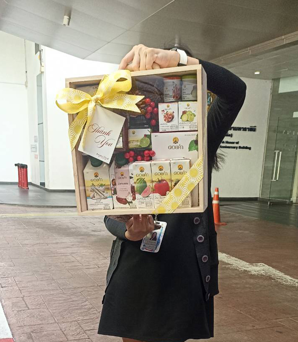 A person holding a wooden gift box decorated with a yellow ribbon. The box contains multiple small boxes with colorful packaging and an assortment of items including artificial berries. The person is wearing a black outfit and standing outside near a building.