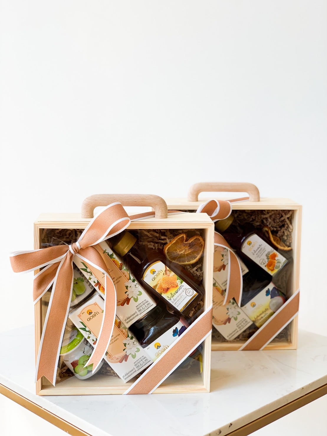 Two wooden gift boxes with handles and ribbons are filled with various items, including fruit, jars of preserves, and other gourmet products. The boxes sit on a white surface against a white background, each tied with a brown ribbon.
