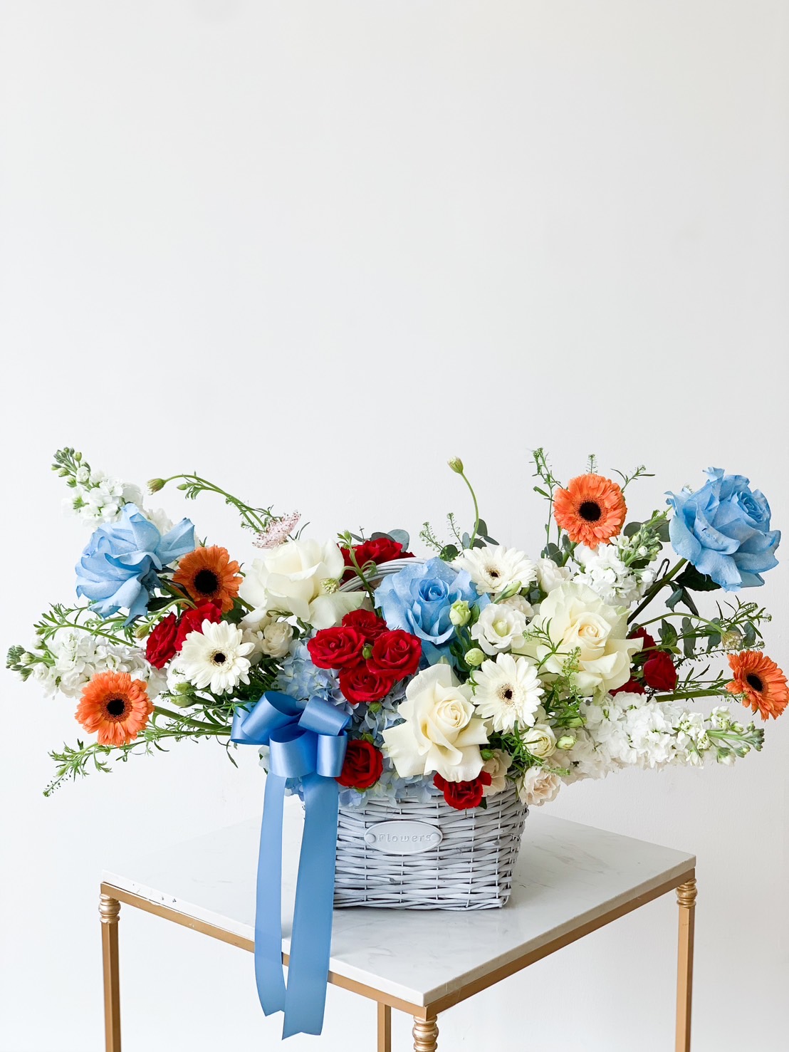A decorative floral arrangement in a white woven basket displayed on a small white table. The Love's Blue Flowers Gift Basket contains light blue roses, red roses, white roses, orange gerberas, white daisies, and greenery, all tied with a blue ribbon. The background is a plain white wall.