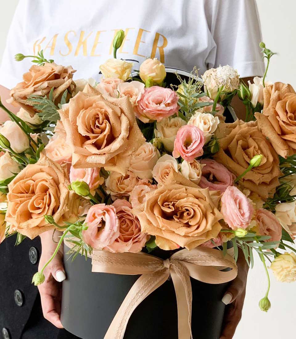 A person adorned in an elegant white shirt cradles a luxurious round, black hatbox from Basketeer, overflowing with an opulent bouquet. The exquisite arrangement features grand brown roses complemented by delicate pink and white blooms, all elegantly tied with a sumptuous beige ribbon showcasing the Basketeer logo.