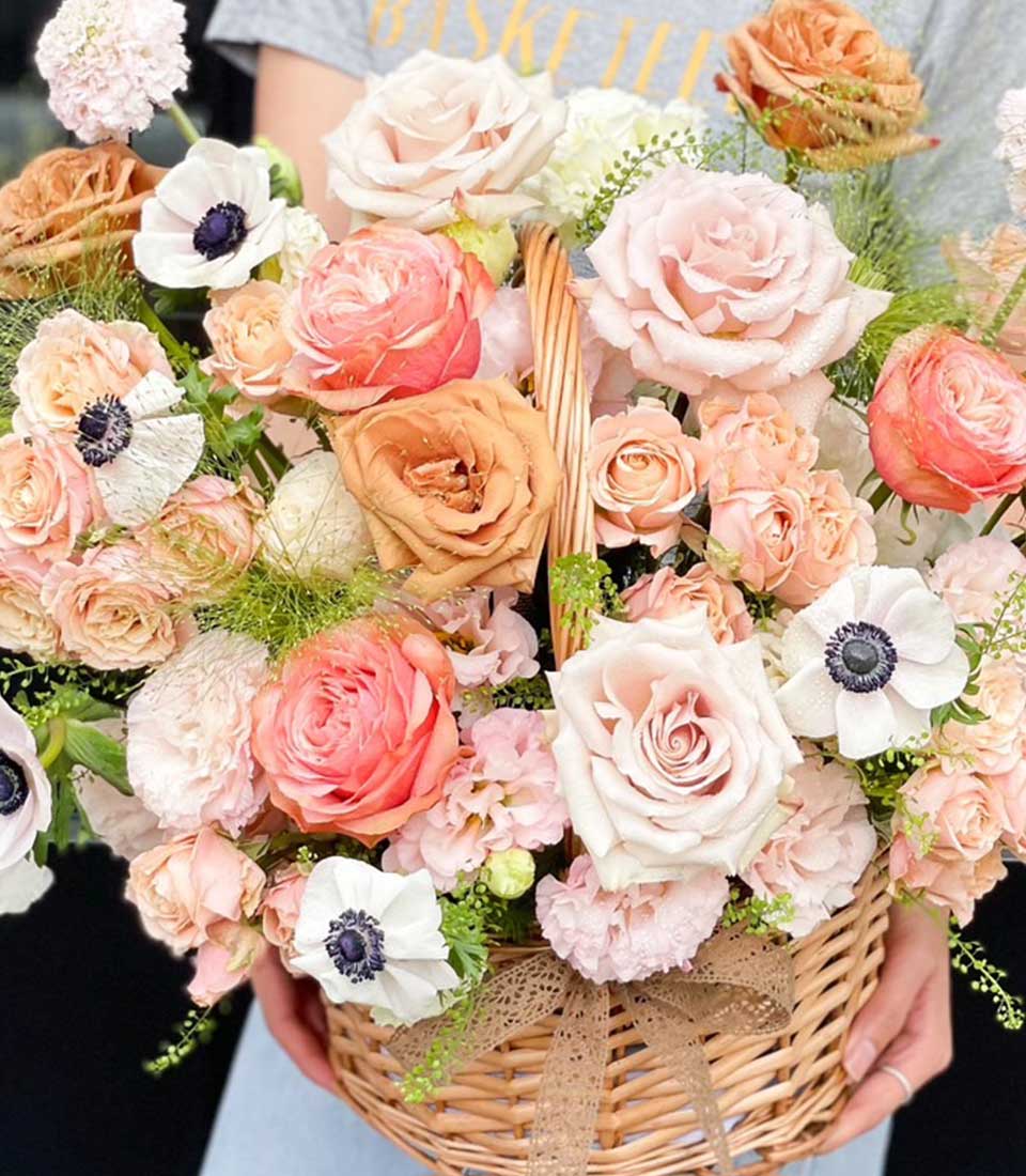 A elegantly manicured hand cradles a luxurious Basketeer wicker basket, brimming with an opulent array of blooming roses and delicate flowers in enchanting shades of peach, pink, and cream. The sumptuous blossoms are interwoven with lush green foliage and neatly tied together with an exquisite lace bow. A glimpse of the sophisticated Basketeer logo on a ribbon adds a touch of exclusivity to this captivating floral arrangement.