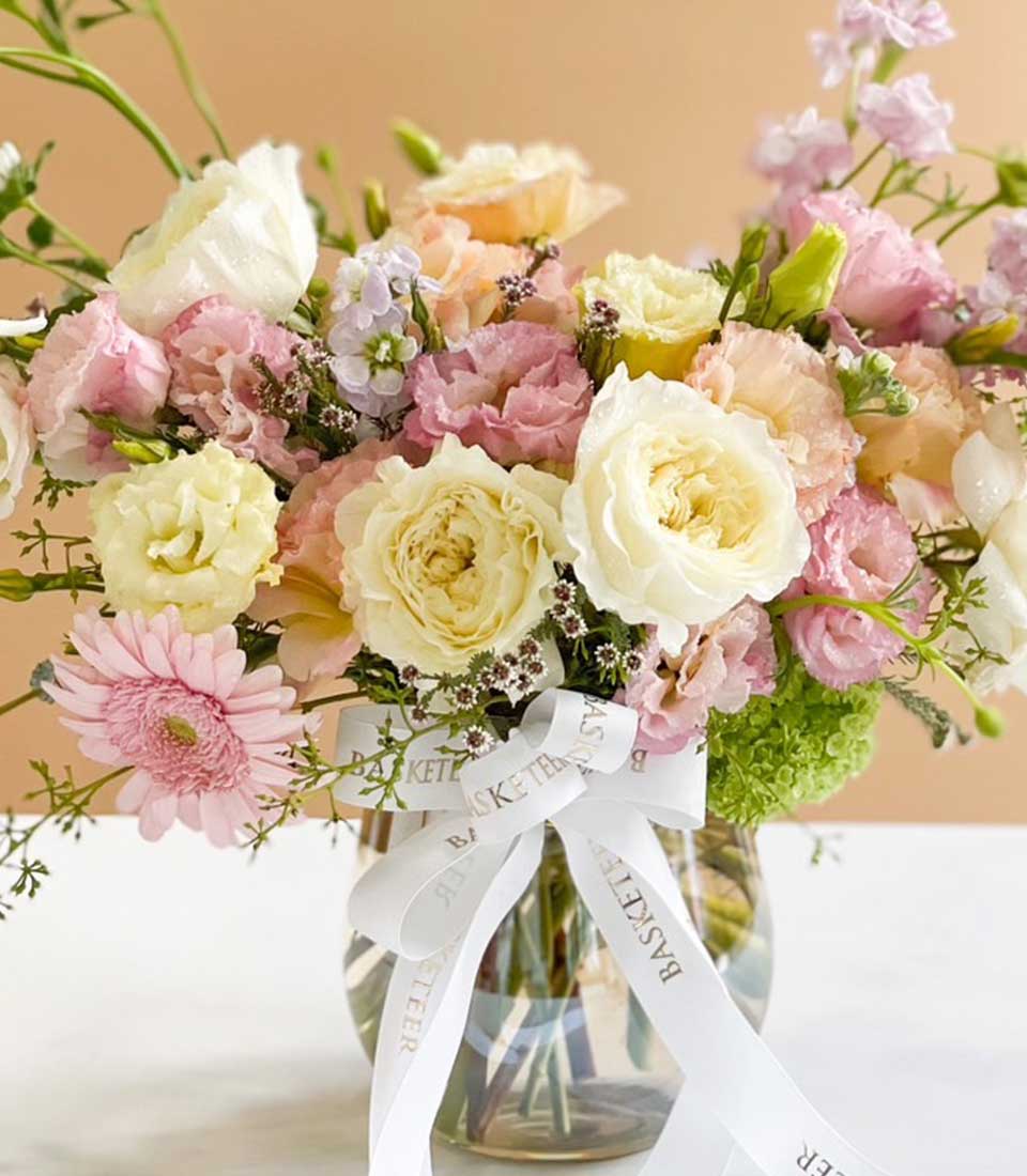 A luxurious floral arrangement in a pristine glass vase, featuring elegant white and blush pink roses intertwined with other soft pastel blooms and lush greenery. The vase is beautifully adorned with a chic white ribbon that proudly displays the 