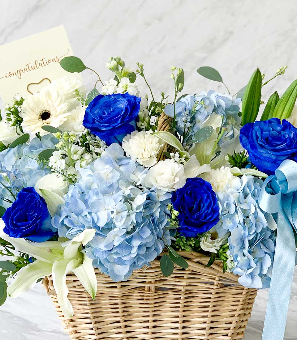 A luxurious wicker basket showcases an exquisite arrangement of flowers, featuring opulent blue roses, delicate light blue hydrangeas, pristine white lilies, and elegant white carnations. A beautifully crafted card with the inscription 