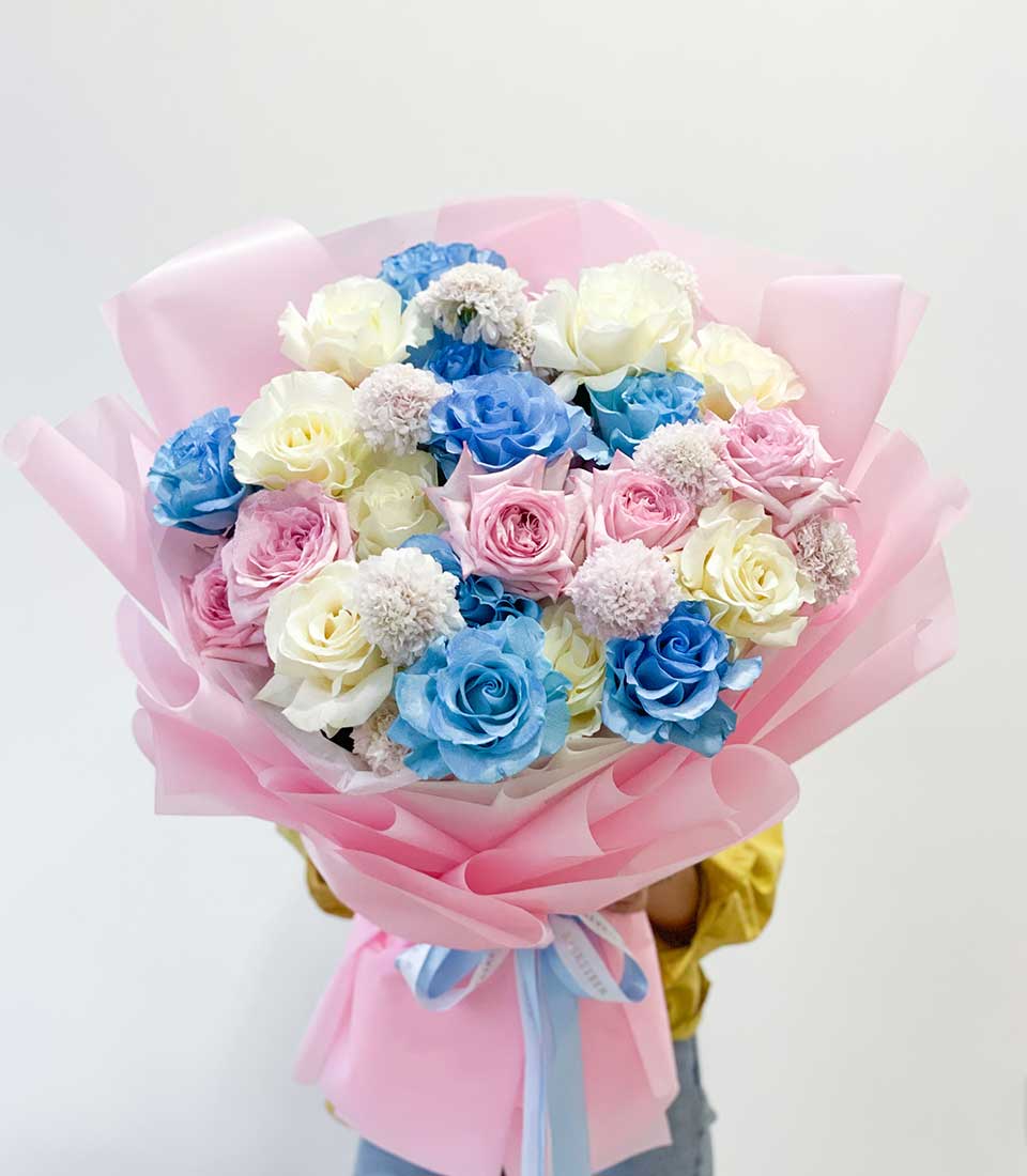 A person elegantly dressed in a yellow top presents the luxurious Sweet Radiant Roses Bouquet from Basketeer, artfully wrapped in exquisite pink paper. This enchanting arrangement showcases a captivating mix of blue, white, and pink roses, delicately accented with petite white blossoms. The bouquet is beautifully tied with a sophisticated blue and white ribbon that exudes elegance. All set against a pristine white backdrop, this scene perfectly highlights Basketeer's commitment to timeless beauty and refinement.