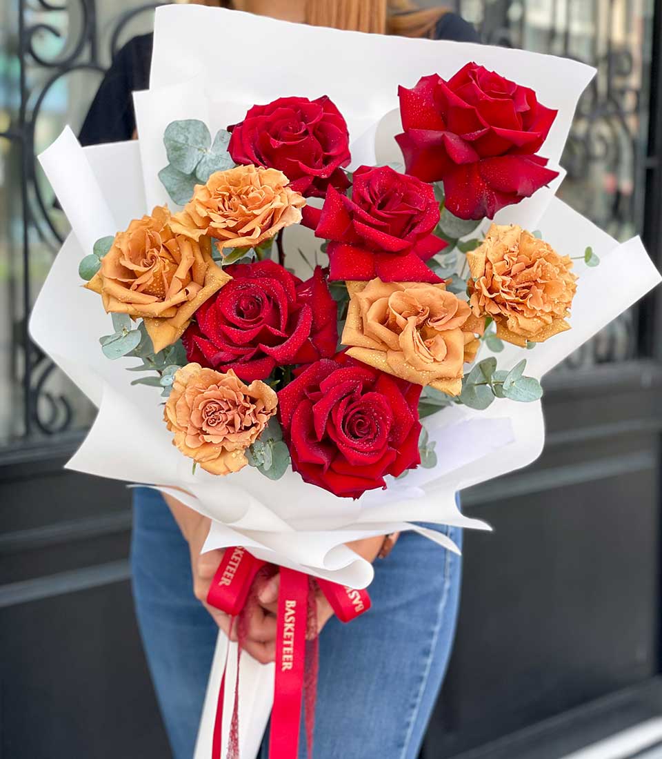 A person cradles an exquisite bouquet featuring six opulent red roses and three delicate peach-hued roses, all elegantly wrapped in pristine white paper adorned with a luxurious red ribbon. Verdant green leaves provide a lush contrast. The individual, dressed in a sophisticated long-sleeve top paired with classic denim jeans, showcases just their hands and lower torso as they present this stunning floral arrangement. A hint of the Basketeer brand's distinguished logo can be seen peeking from the ribbon, adding an extra touch of elegance to the scene.