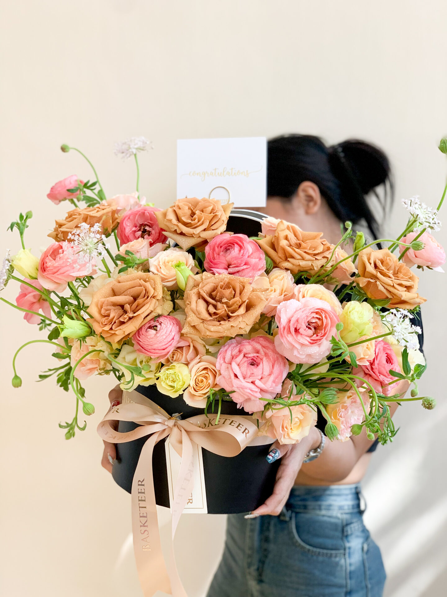 A stylish individual clad in a pristine white t-shirt and an elegant black skirt cradles a luxurious black circular hatbox adorned with the Basketeer logo. This exquisite arrangement features a stunning selection of peach and pink roses, complemented by lush greenery, all gracefully tied together with a sophisticated beige ribbon.