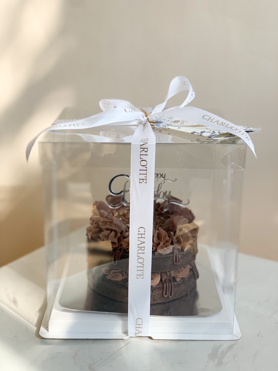 A decadent chocolate cake with rich swirls and a “Happy Birthday” topper, presented in a CHARMOTTE ribboned box, Charlotte style.