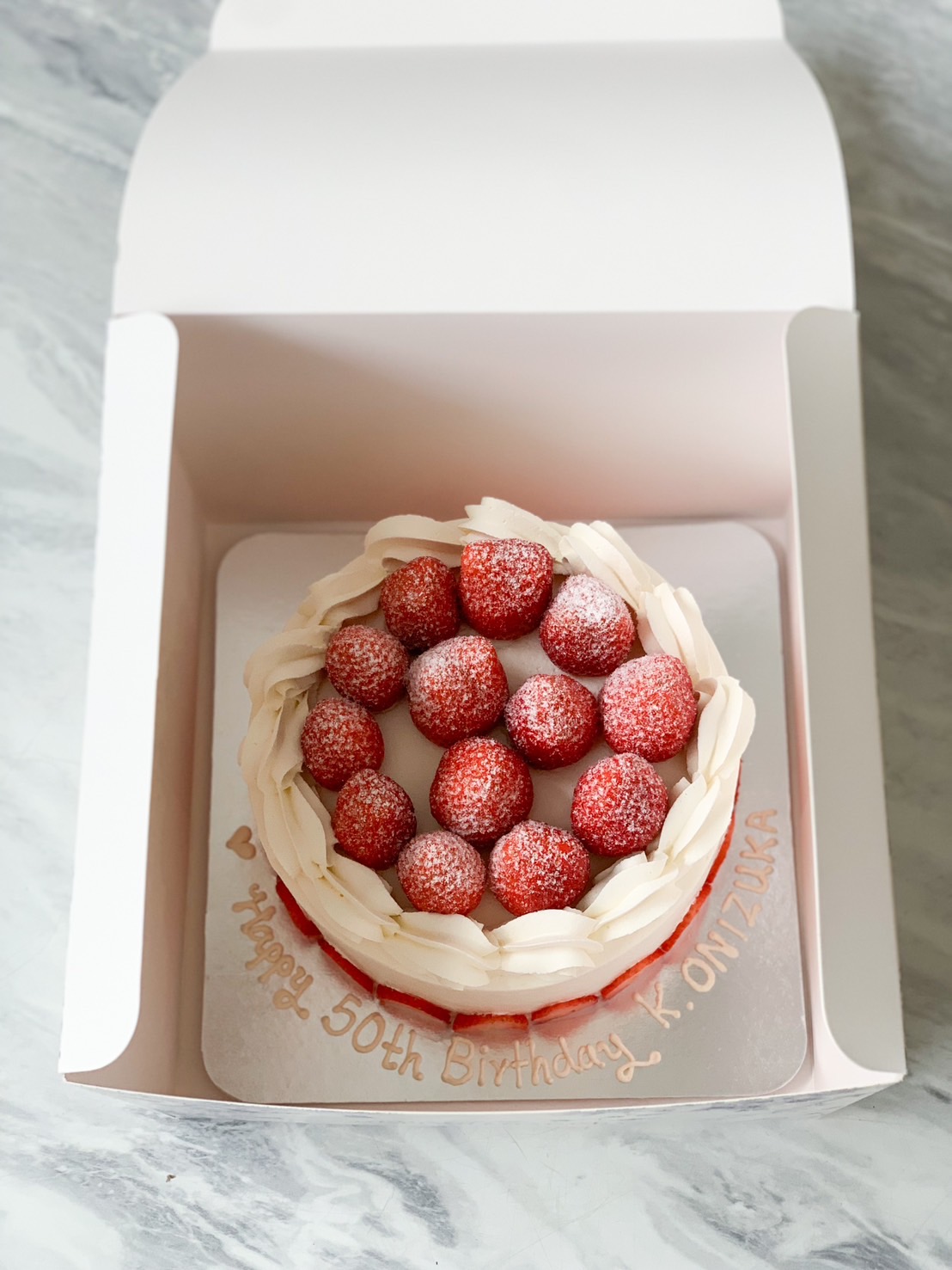 Indulge in the epitome of luxury with Basketeer's strawberry lover's dream cake. This exquisite birthday cream cake is elegantly presented on a marble stand with a refined wooden base. Adorned with whole strawberries delicately dusted in powdered sugar and artfully arranged slices around the bottom, it's a masterpiece for both the eyes and palate. A resplendent gold 