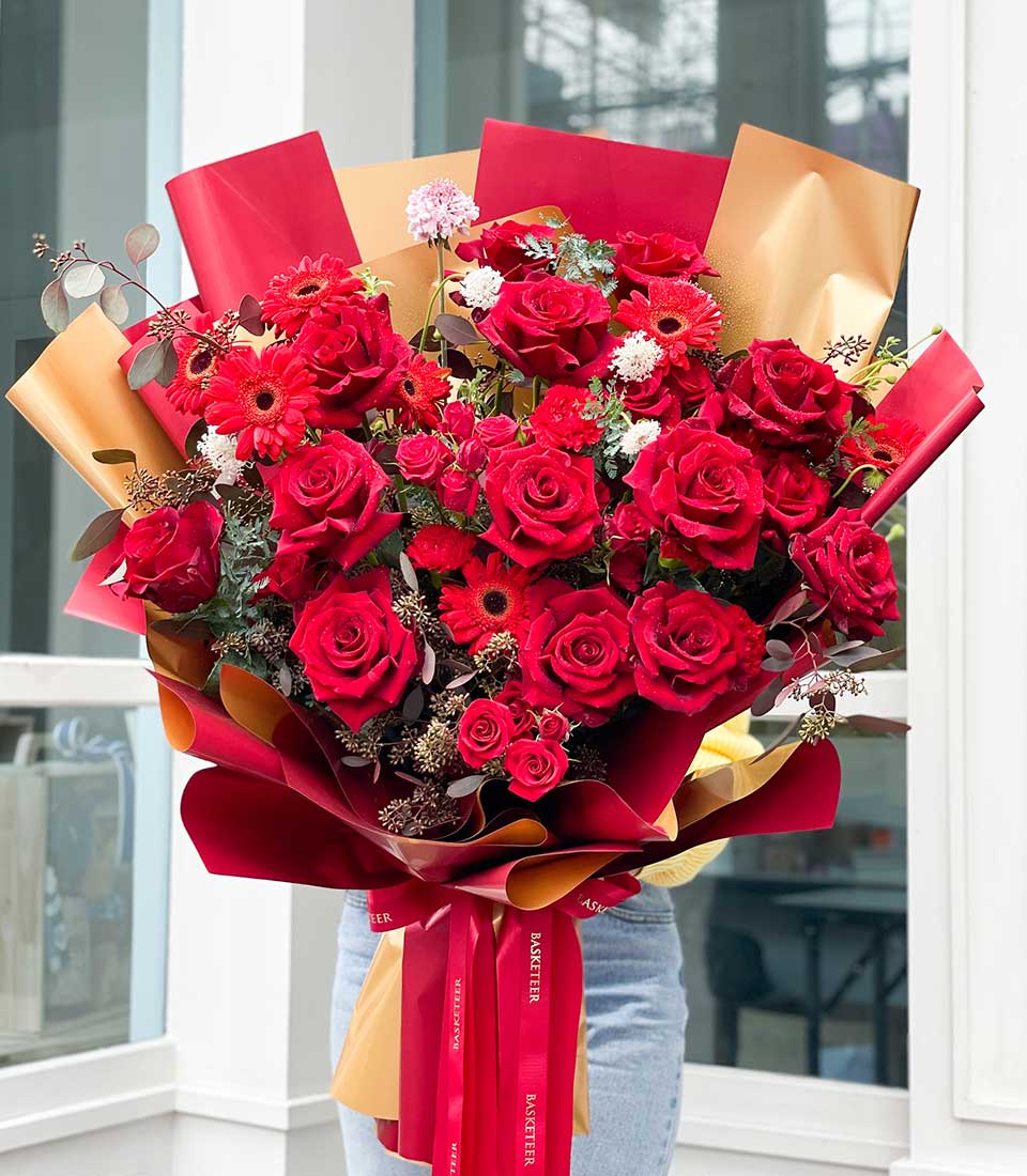 A person delicately holds an opulent bouquet brimming with vibrant red roses, striking red gerbera daisies, and lush assorted greenery. The luxurious Sizzling Crimson Affection Bouquet is elegantly wrapped in rich red and golden paper, secured with a sumptuous red ribbon bearing the exquisite Basketeer logo. The scene exudes sophistication and charm, making it a magnificent floral arrangement perfect for any grand occasion.