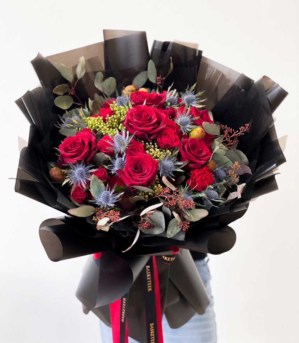 A person is elegantly holding a luxurious bouquet of vibrant red roses, striking blue thistles, and rich greenery wrapped in sophisticated black paper adorned with opulent red ribbons. This stunning arrangement from Basketeer showcases a perfect blend of bold flowers and lush foliage, exuding unparalleled elegance and charm. The Basketeer branding subtly graces the ribbon, enhancing the bouquet's allure and refinement.