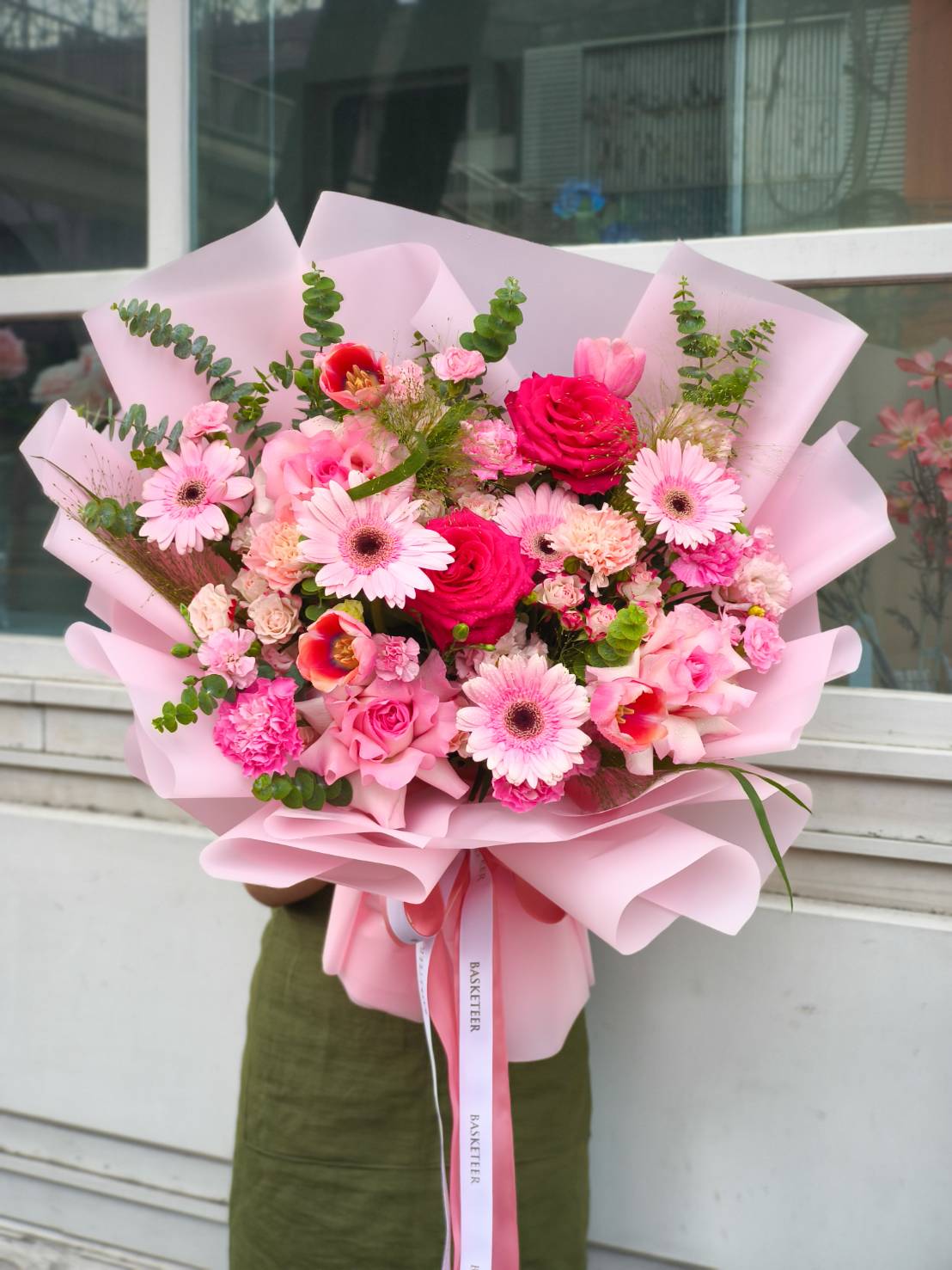 Large bouquet with vibrant pink roses, gerberas, carnations, tulips, and lush greenery, wrapped in pink paper.