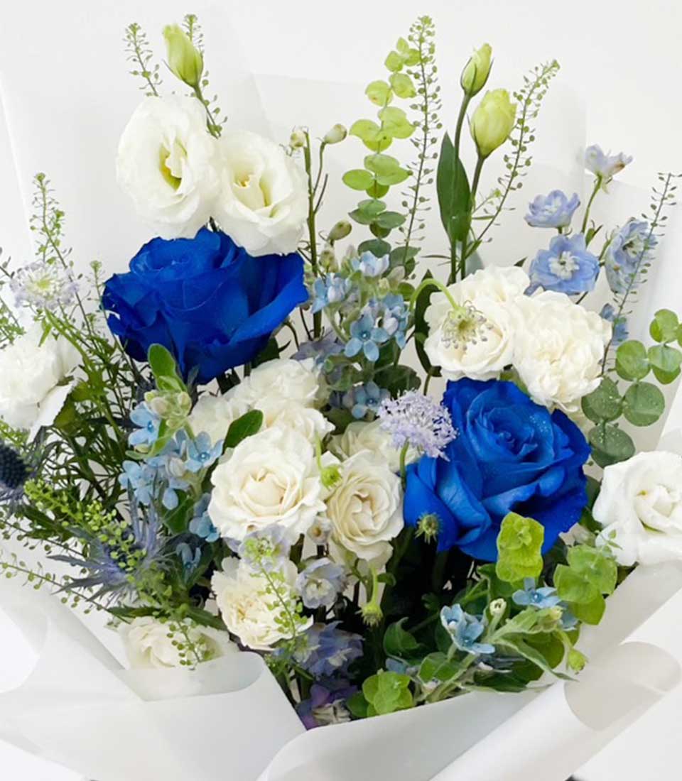 A stunning bouquet of blue and white flowers, artistically arranged with lush greenery and elegantly wrapped in white paper with a branded ribbon.