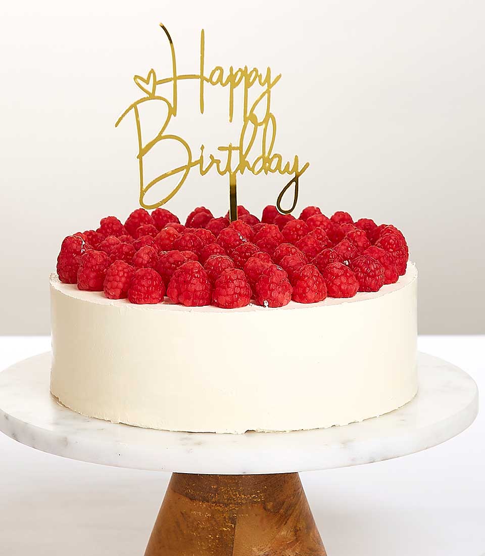 A decadent white-frosted Temptation Cake adorned with luscious, plump raspberries graces an opulent marble cake stand with a luxurious gold base. Atop the cake, a sophisticated gold topper elegantly spells 