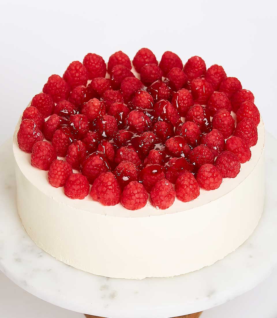 A sumptuous round Temptation Cake, elegantly adorned with luscious raspberries—some artfully glazed to a glistening perfection. This luxurious creation is meticulously presented on a pristine white marble cake stand, embodying sophistication. Against the minimalist white backdrop, the Basketeer-branded ribbon whispers hints of exclusivity and grandeur. The smooth surface of the cake is flawlessly crowned with an even layer of vibrant raspberries, inviting pure indulgence.