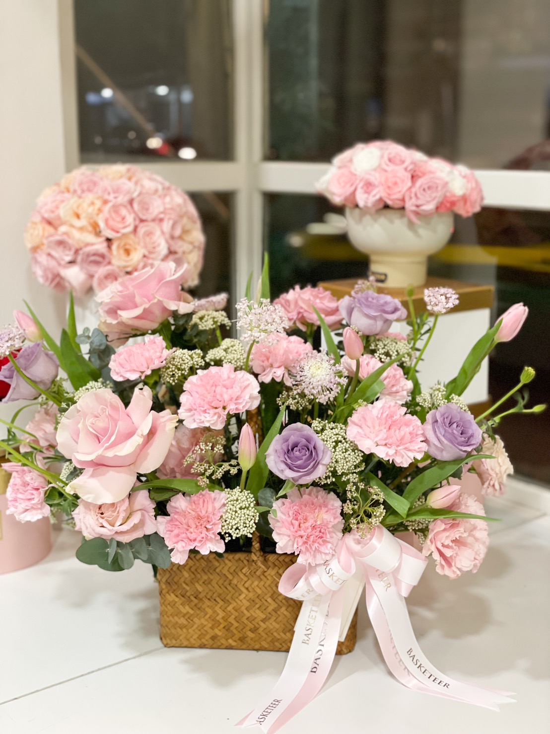A luxurious pink and lavender floral arrangement in a woven basket, featuring roses, carnations, and lisianthus, tied with a Basketeer ribbon.