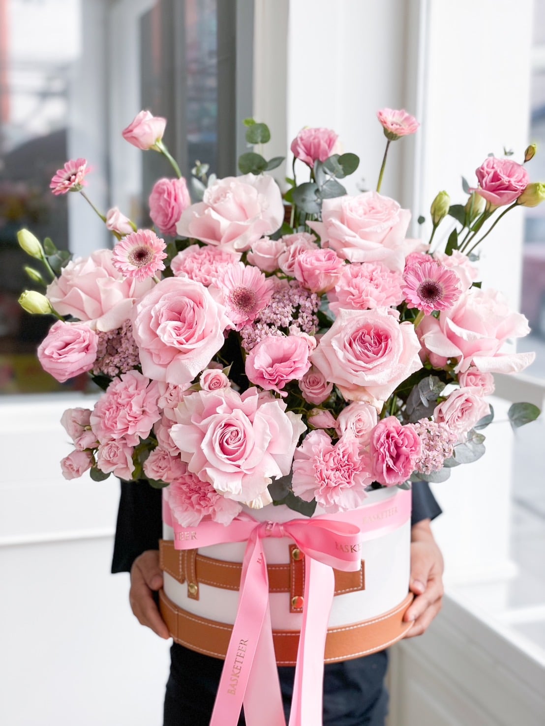 A person holds a large, round hatbox filled with a Sweet Pink Blooming Gift of roses, carnations, and gerberas. The box is adorned with a pink ribbon and bow. The background shows a bright window.