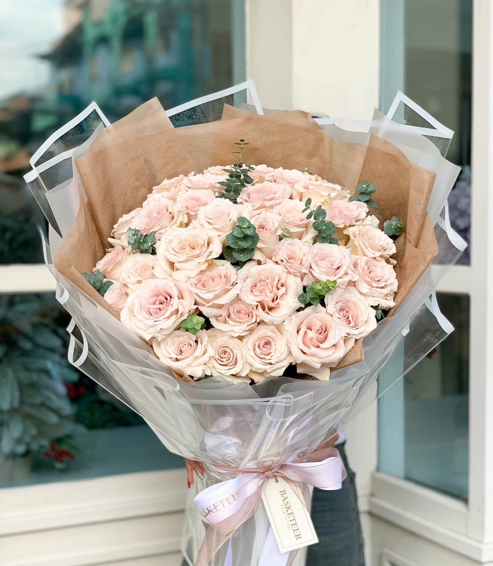 A lavishly opulent bouquet of Pink Mondial Roses, artistically wrapped in luxurious layers of crisp white, delicate pink, and rich brown paper. The arrangement is gracefully adorned with elegant pink and white ribbons, adding a touch of sophistication. Displayed against a pristine white backdrop, this masterpiece embodies the elegance of Basketeer’s signature style. Look closely to find the distinguished Basketeer logo subtly woven into the ribbon and adorning the intricately designed wrapping paper.