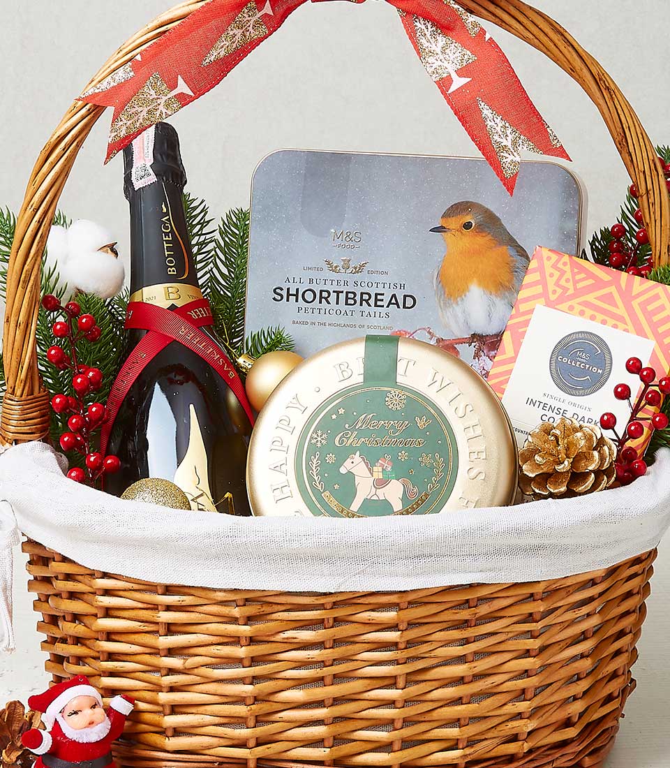 A luxurious wicker basket brimming with holiday delights from Basketeer, elegantly wrapped in a rich red ribbon that features the Basketeer logo. Nestled inside, you'll find a bottle of premium sparkling wine, a beautifully illustrated tin of shortbread cookies adorned with an enchanting robin, and a festive round tin of delectable chocolates. Accents of gold pinecones and vibrant red berries add a touch of opulence, complemented by a charming Santa figurine that completes this inviting seasonal arrangement.