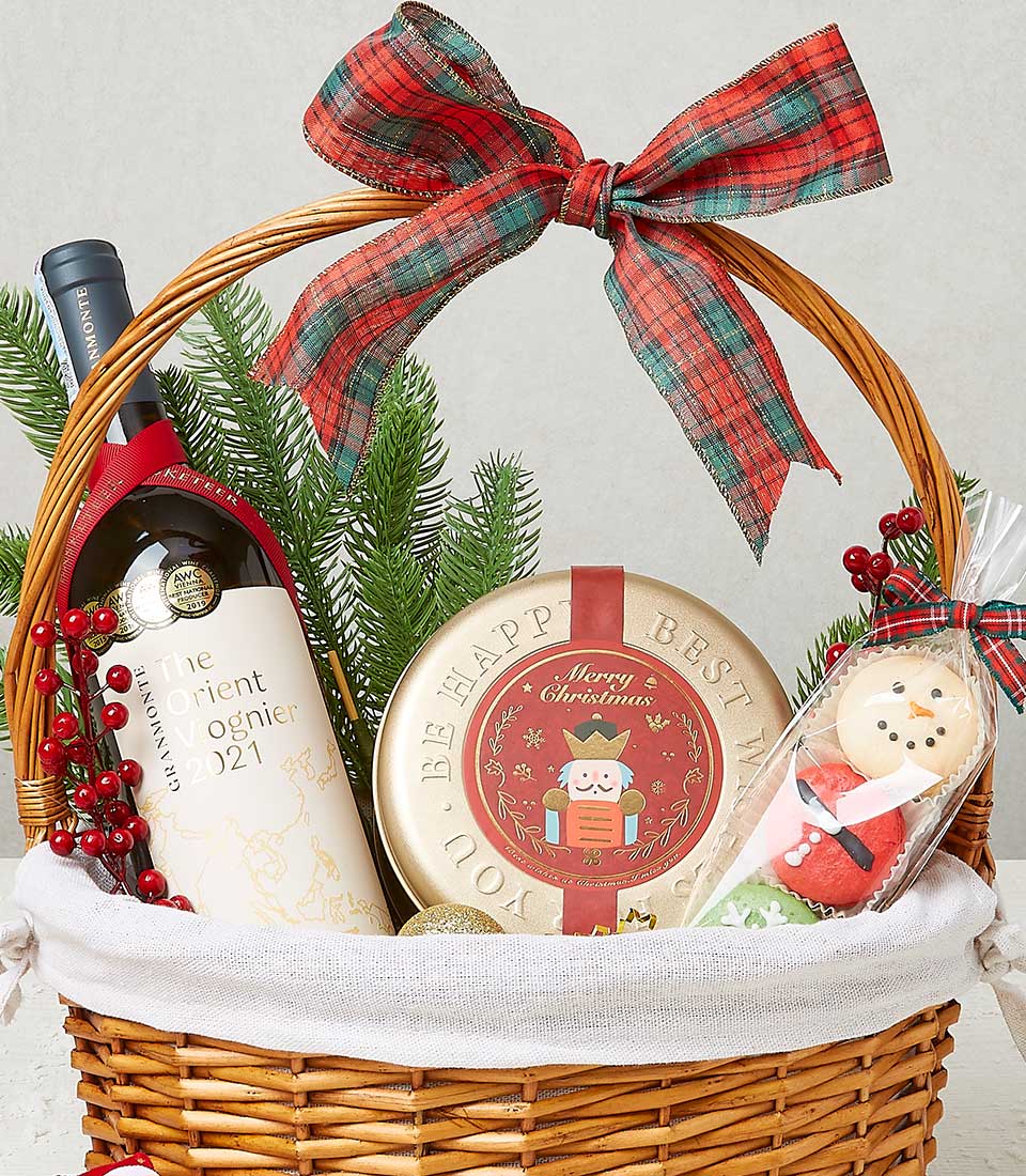 A sumptuous gift basket adorned with a lavish red plaid bow, featuring the distinguished Basketeer logo elegantly displayed. Nestled within this opulent arrangement is a fine bottle of wine, paired perfectly with a tin of gourmet Christmas cookies bearing intricate designs. A sprig of evergreen adorned with vibrant red berries enhances the festive allure. Additionally, a pristine clear bag contains exquisitely decorated Christmas cookies. This luxurious ensemble emanates warmth and holiday cheer through its rich, colorful decorations – an impeccable offering from Basketeer for those who appreciate the finer things in life.