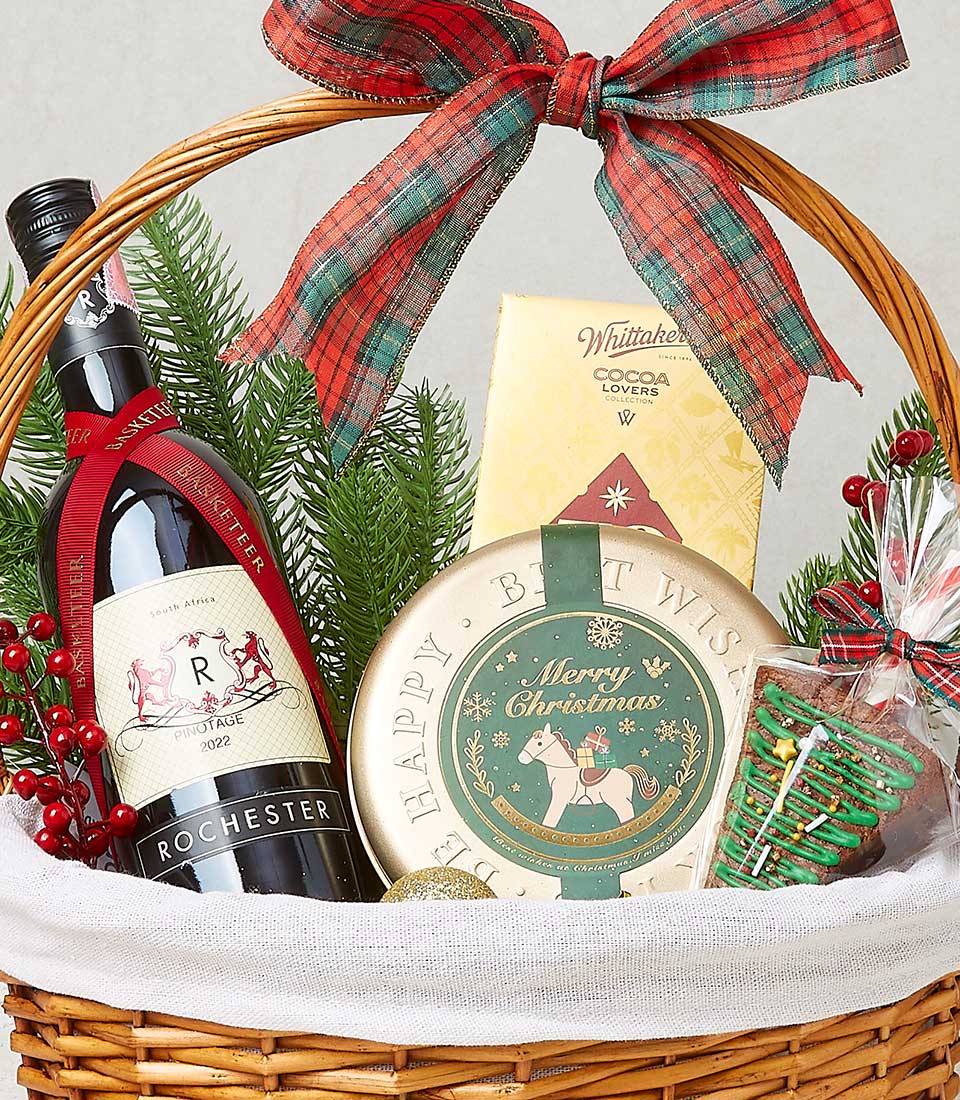 A lavish and inviting holiday scene featuring a deluxe wicker basket adorned with an elegant red and green plaid bow and festive fir branches. Inside, discover a selection of refined indulgences: a distinguished bottle of red wine, a charming tin labeled 