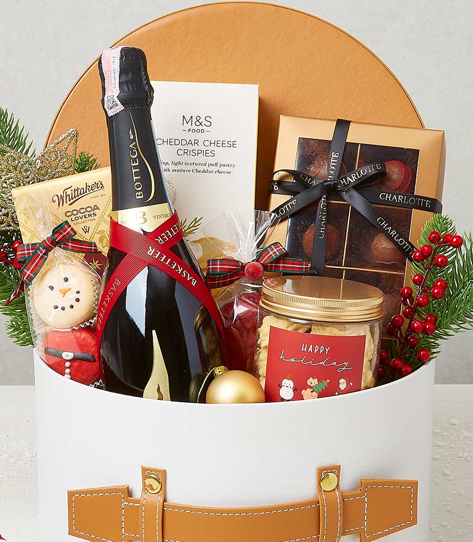 A lavish gift basket from Basketeer brimming with holiday elegance: a bottle of fine sparkling wine, savory cheddar cheese crispies, decadent chocolates, and a charming gingerbread cookie. Accompanied by a box of rich cocoa nibs, assorted candies, and other delectable treats. The basket is beautifully adorned with festive pine branches and vibrant red berries, all meticulously arranged in an exquisite white and brown wicker basket. A luxurious ribbon featuring the Basketeer logo adds the perfect finishing touch to this splendid seasonal offering.