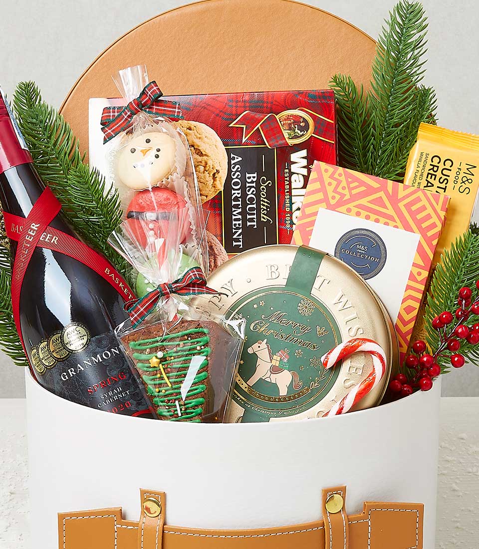 A luxurious Basketeer gift basket beautifully presented with an exquisite bottle of red wine, a selection of gourmet snacks, a decadent box of assorted chocolates, and a tin of buttery biscuits. Nestled among holiday greenery adorned with vibrant red berries are delightful treats including a festive red and green lollipop, an artfully decorated Christmas tree cookie, and a premium yellow snack bar. The scene is elegantly wrapped with the Basketeer ribbon, exuding sophistication and inviting indulgence.