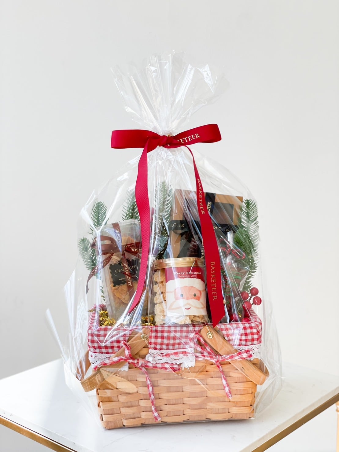 A wicker gift basket wrapped in clear cellophane, tied with a red ribbon. The basket features festive items like a Santa-themed container, chocolates, greenery, and decorative elements with a red and white checkered cloth at the base.